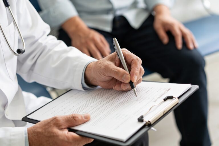 Doctor in a white coat writes on a clipboard with a pen; a stethoscope rests nearby as a patient sits in the background.