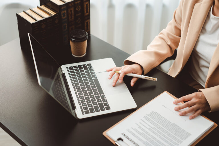 Businesswoman's hands typing on a laptop and holding a pen while reviewing legal documents and agreements on a clipboard, managing important professional tasks in a modern office environment